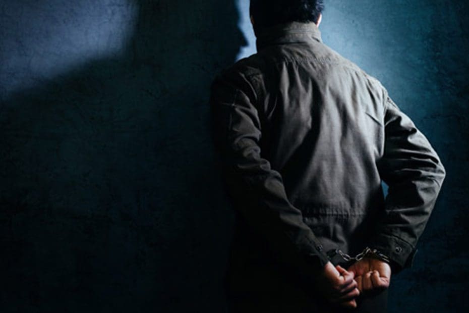dark image of a man staring at a wall with his hands handcuffed behind his back