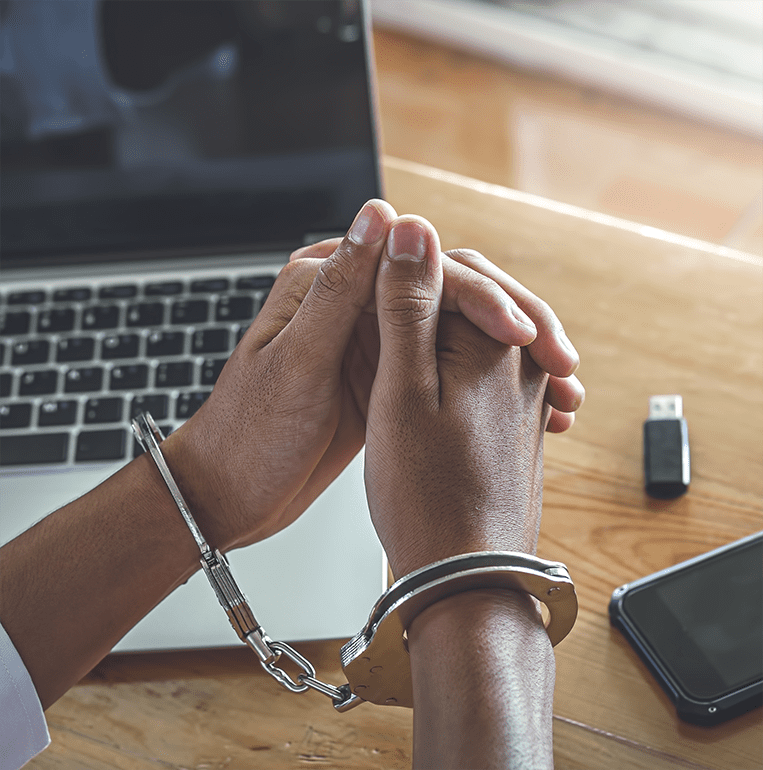 Two hands of person arrested with handcuffs in front of a laptop and a cell phone